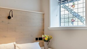 Ein Schlafzimmer mit einem hölzernen Kopfende, einem Nachttisch mit Blumenvase und einem Fenster mit Buntglasdekor.