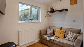 A cozy living room with a sofa, a radiator, a window with a view of greenery, and a hanging shelf with a vase.