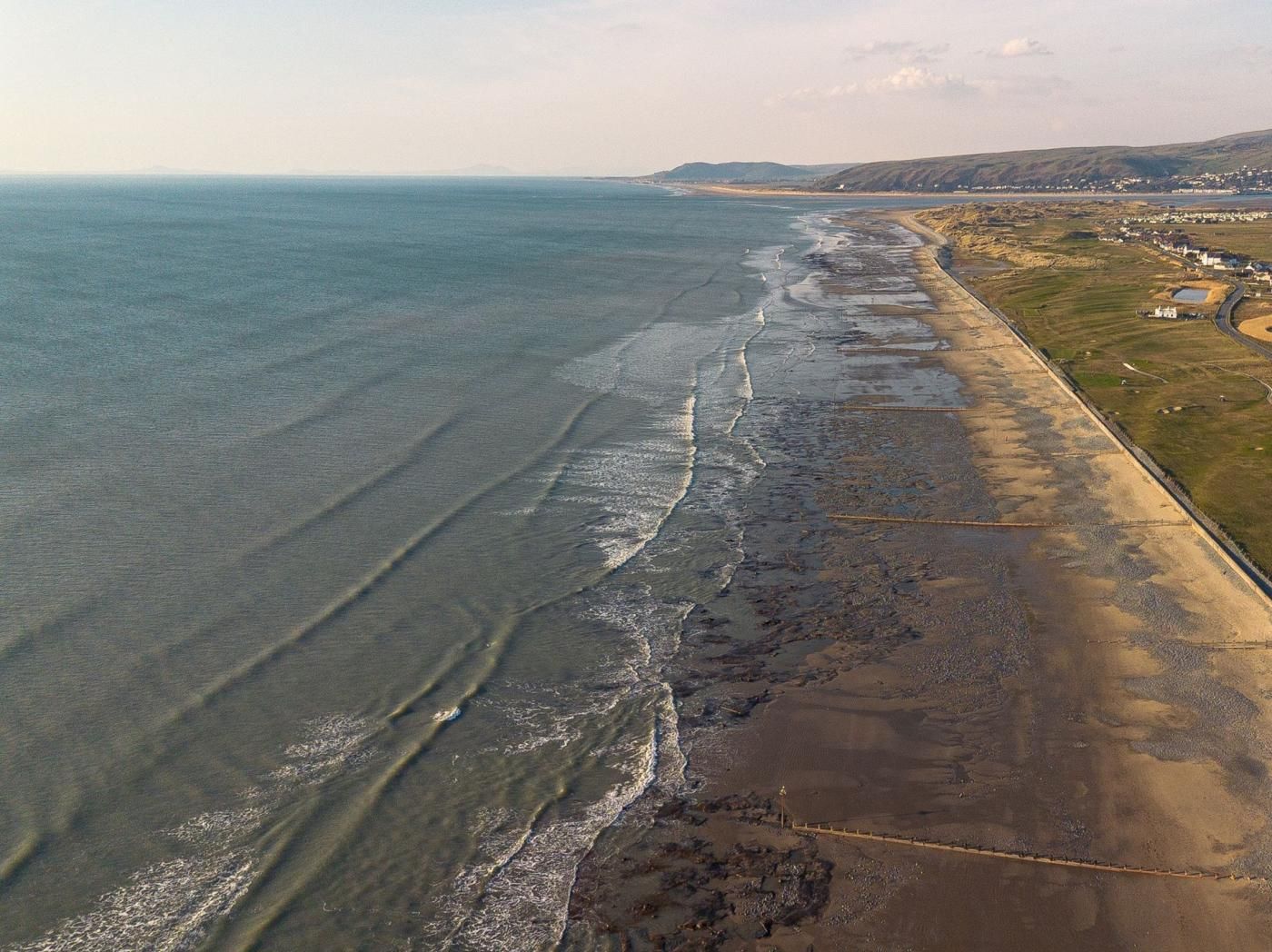 An aerial view of a coastal area with a sandy beach, a clear demarcation of land and sea, and a distant town.