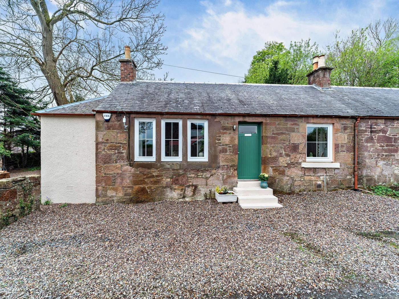 A stone cottage with a green door, three windows, and a gravel driveway.