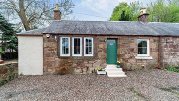 A stone cottage with a green door, three windows, and a gravel driveway.