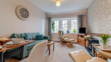 A living room with a sofa, a chair, a dining table with a bowl and a wine glass, a television, and a clock on the wall.