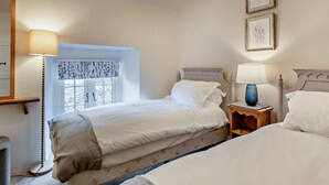 Une chambre avec deux lits, une table de chevet avec une lampe, une fenêtre ornée de rideaux à motifs floraux et des œuvres d’art encadrées accrochées au mur.
