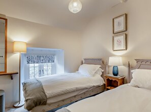 Ein Schlafzimmer mit zwei Betten, einem Nachttisch mit Lampe, einem Fenster mit blumigen Vorhängen und gerahmten Bildern an der Wand.