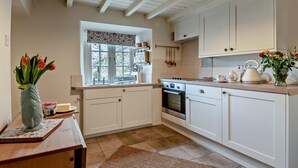 Une cuisine avec des meubles blancs, une cuisinière, un évier et une fenêtre ornée d’un rideau à motifs floraux.