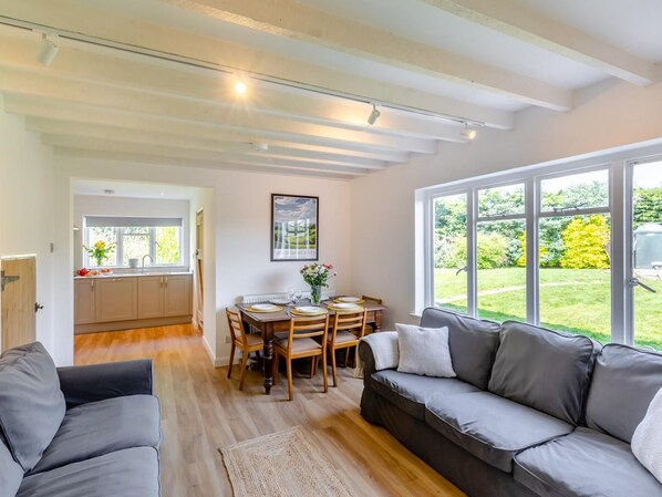 A modern living room with a sofa, a dining table with chairs, and a kitchen area with cabinets and a window.