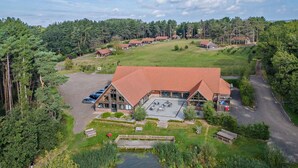 A lodge with a large parking area, a pond, and a forested backdrop. - 3 Bed in King's Lynn (oc-96035) (Middleton)