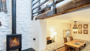 A cozy room with a wood-burning stove, a dining table, chairs, a lamp, and framed pictures on the wall. - 1 Bed in Tavistock (oc-96009) (Cudlipptown)