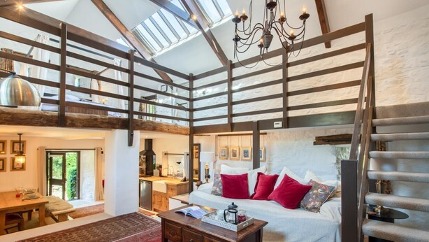A spacious loft-style room with a high ceiling, exposed wooden beams, a large bed with red pillows, a dining area with a table and chairs, and a view of the outdoors through skylights. - 1 Bed in Tavistock (oc-96009) (Cudlipptown)