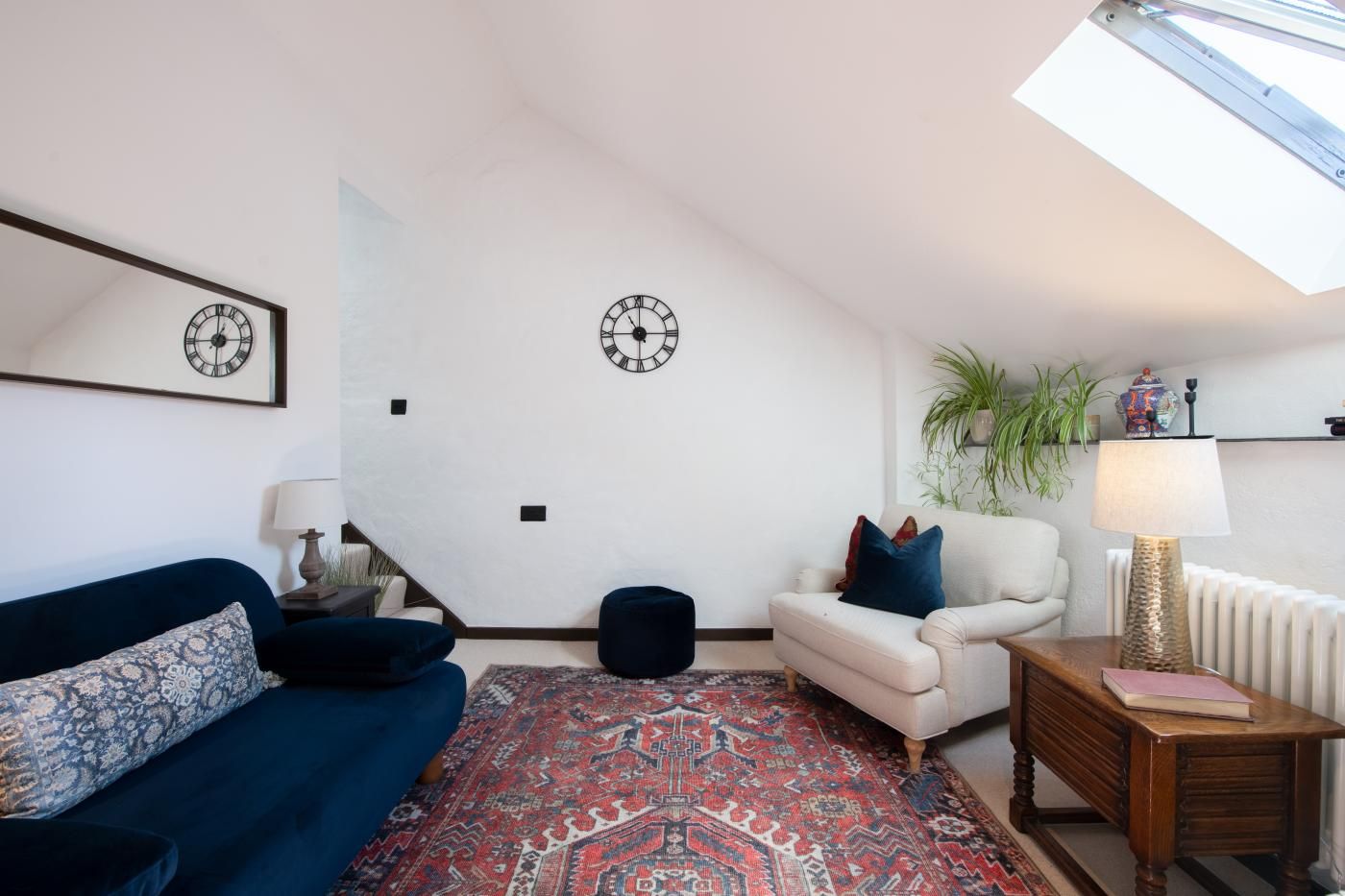 A living room with a blue sofa, a white armchair, a patterned rug, and two wall clocks.