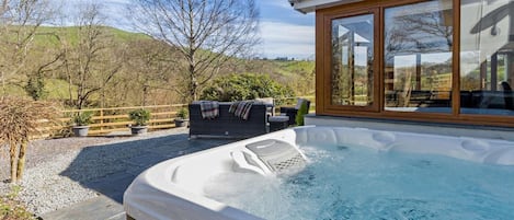 A hot tub with a wooden deck, a seating area with a table and chairs, and a view of the surrounding landscape.