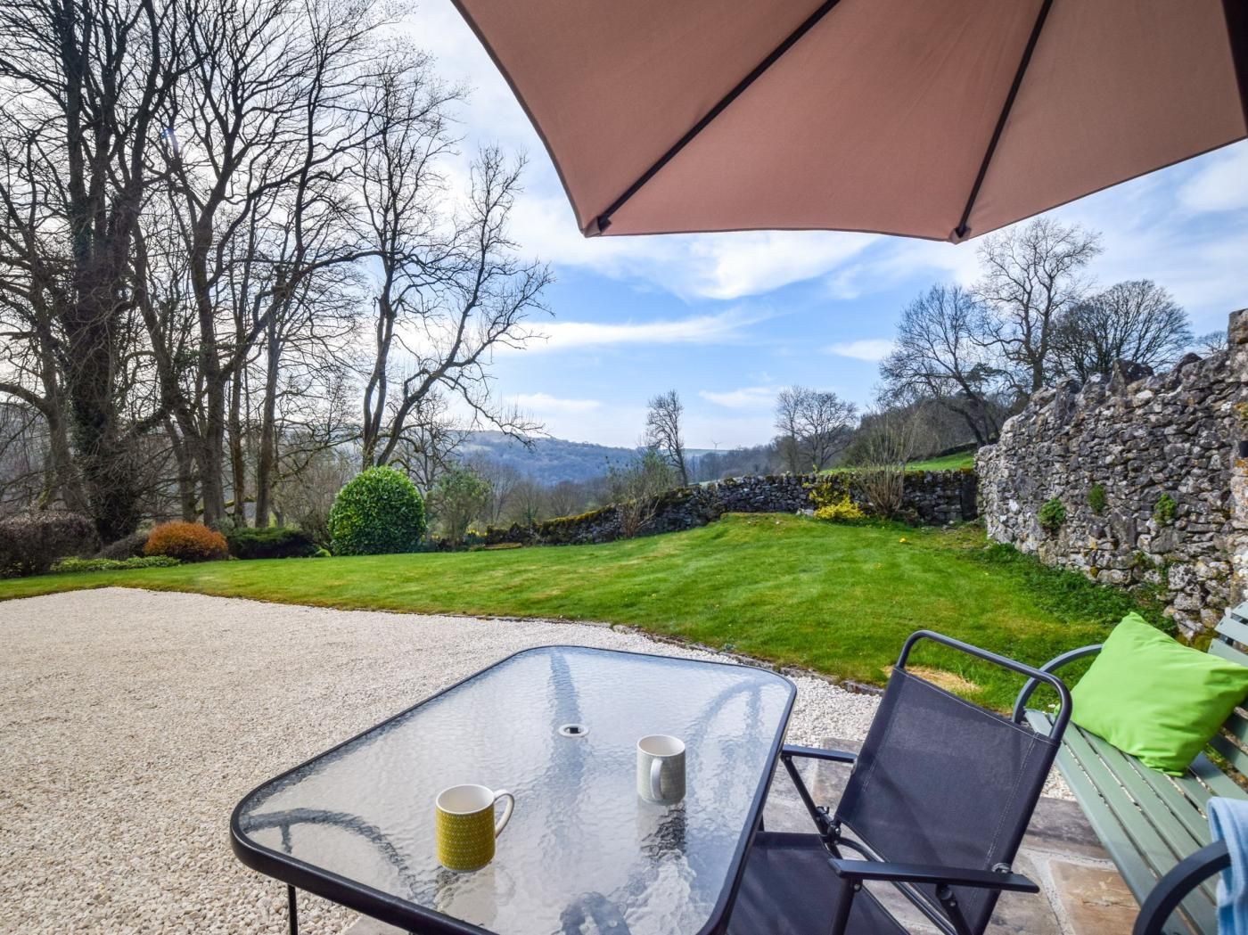 An outdoor patio with a glass table, two chairs, a table lamp, and a large umbrella.