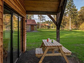 Ein Holzhaus mit einer überdachten Terrasse, einem Picknicktisch und Blick auf eine Rasenfläche und Bäume.
