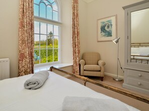 A bedroom with a large bed, a chair, a desk, a lamp, and a window with curtains. - 2 Bed in Beaminster (oc-78567) (Beaminster)