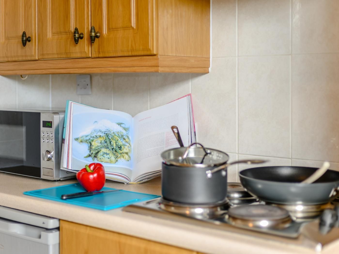 Une cuisine avec des meubles de rangement en bois, un four à micro-ondes, un livre, un poivron rouge, une casserole et une poêle sur un plan de travail.