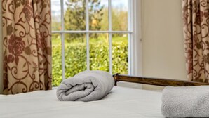 A neatly made bed with a rolled towel and a wooden bedpost in a room with patterned curtains and a window with a view of greenery. - 2 Bed in Beaminster (oc-78567) (Beaminster)