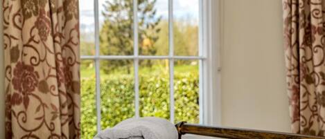 Una cama bien hecha con una toalla enrollada y un pilar de madera en una habitación con cortinas estampadas y una ventana con vistas a la vegetación.