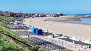 Eine Küstenstraße mit einem Strand zur Rechten, blauem Himmel und Gebäuden im Hintergrund.