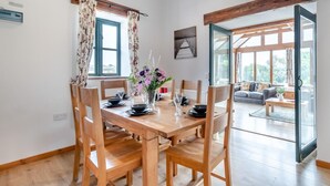 A dining area with a wooden table, chairs, and a vase of flowers. There is a window with curtains, a painting on the wall, and a view of a living room with a sofa and a coffee table. - 3 Bed in Pembroke (oc-44349) (Hundleton)