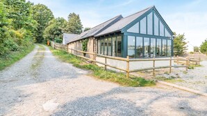 A building with a sloped roof and large windows, surrounded by a gravel path and wooden fence. - 3 Bed in Pembroke (oc-44349) (Hundleton)