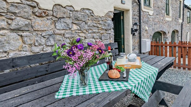 Un coin repas extérieur rustique, avec une nappe à carreaux, un vase de fleurs et un pain sur un banc en bois.