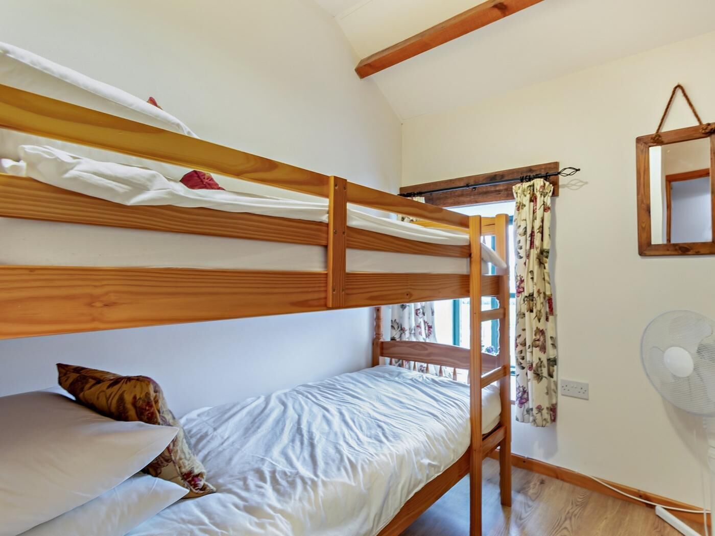 Ein Schlafraum mit Etagenbetten, Holzboden, einem kleinen Fenster mit blumigen Vorhängen und einem Wandventilator.
