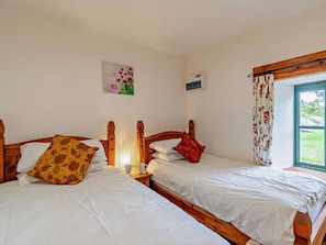 A room with two wooden beds, a window with floral curtains, a wall-mounted air conditioning unit, and a small table with a cup. - 4 Bed in Pembroke (oc-44351) (Hundleton)