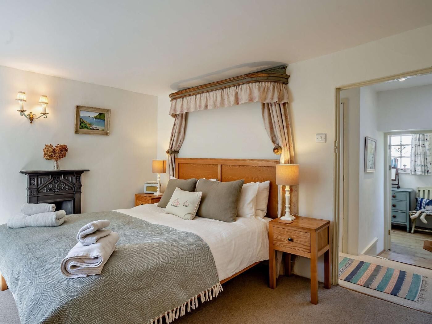 A bedroom with a bed, bedside tables, a nightstand, a fireplace, and a view into another room.
