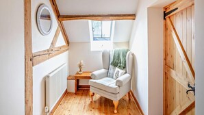 Een gezellig zolderkamertje met houten balkenplafond, een witte radiator, een klein tafeltje met een vaas, een witte fauteuil en uitzicht door een raam.