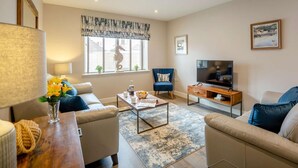 A living room with a sofa, armchair, coffee table, TV, and a window with a seahorse decoration. - 3 Bed in Seahouses (oc-81486) (Seahouses)