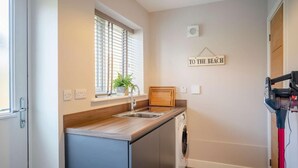 A compact kitchen with a built-in washing machine, a window with blinds, and a sign on the wall. - 3 Bed in Seahouses (oc-81486) (Seahouses)