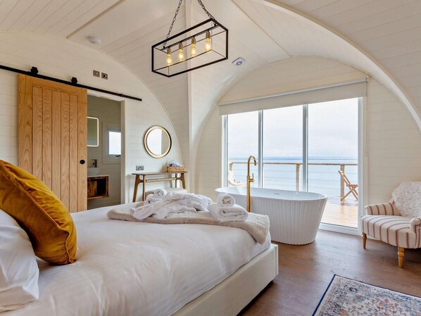 A bedroom with a large bed, a bathtub, a chair, and a view of the sea.
