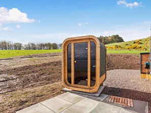 A wooden sauna structure with a glass door, set on a concrete platform in a natural landscape.