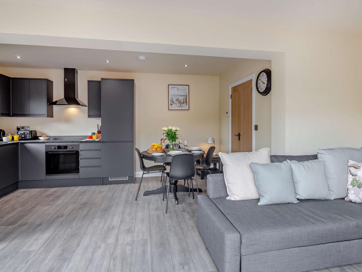 A modern kitchen with dark cabinetry, a dining area with a table and chairs, and a grey sofa with multiple cushions.