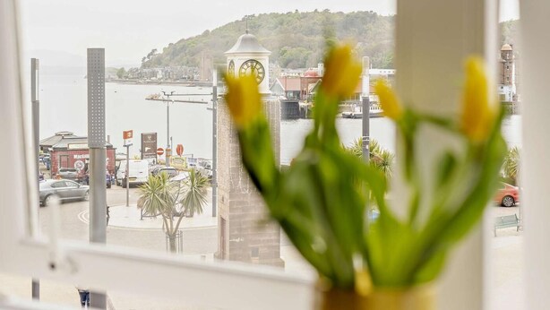 A view of a clock tower, a body of water, and a cityscape through a window with a vase of flowers in the foreground. - 1 Bed in Oban (oc-96079) (Oban)