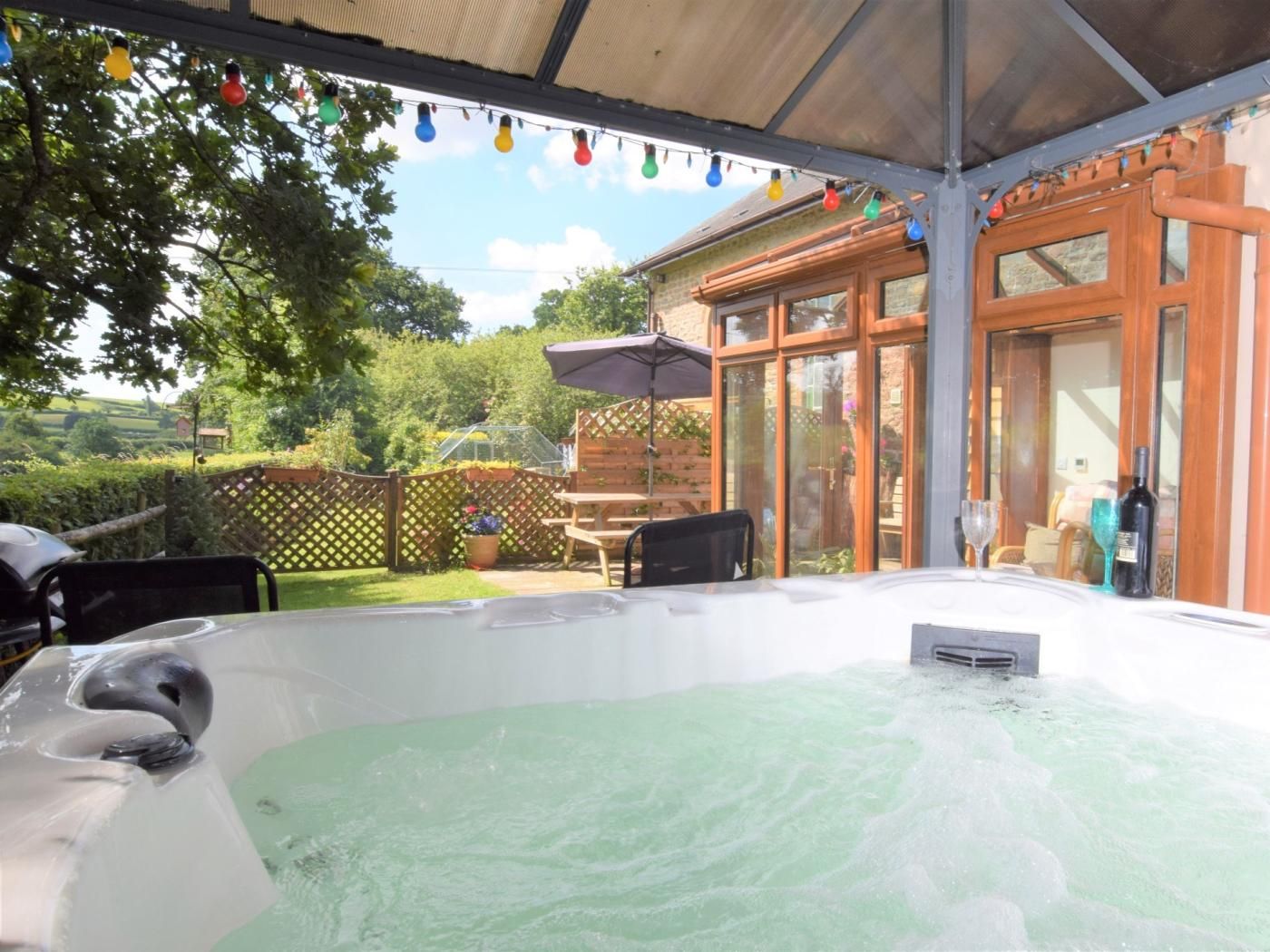 Un bain à remous installé sous une terrasse couverte, avec une clôture en bois et une porte vitrée en arrière-plan.