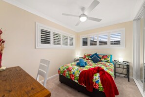 A bedroom with a bed, a ceiling fan, a window with blinds, a bedside table, and a chair.