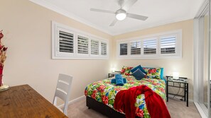 Ein Schlafzimmer mit einem Bett, Deckenventilator, zwei Fenstern mit Fensterläden, einem Nachttisch und einem Sessel.