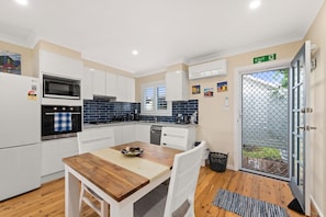 A modern kitchen with white cabinets, a microwave, an oven, and a sink. There's a dining table with a bowl of fruit and a rug.