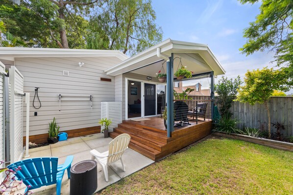 A residential backyard with a covered patio, a barbecue grill, a white chair, and a blue chair.