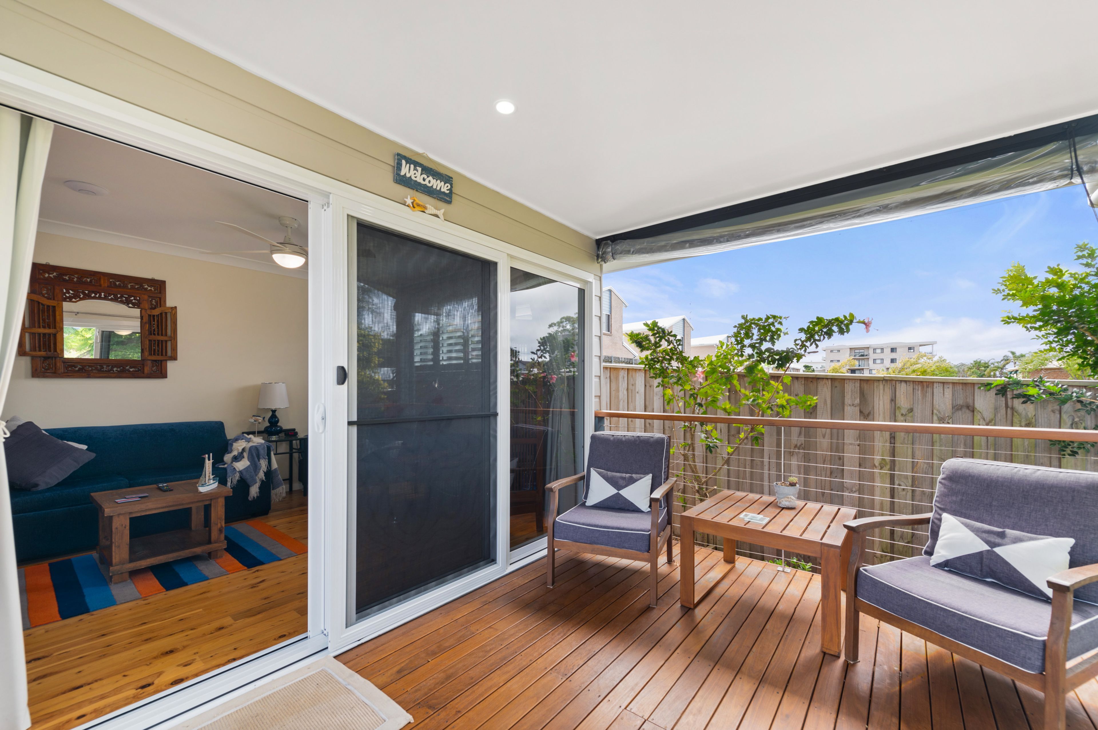 A covered outdoor deck with a sliding glass door leading to a living room, furnished with a sofa, a coffee table, and two chairs.