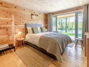 A bedroom with a wooden ceiling, a bed with a quilt, a nightstand, a lamp, and a view of a lake through large windows. - 3 Bed in Cullompton (oc-96118) (Willand)