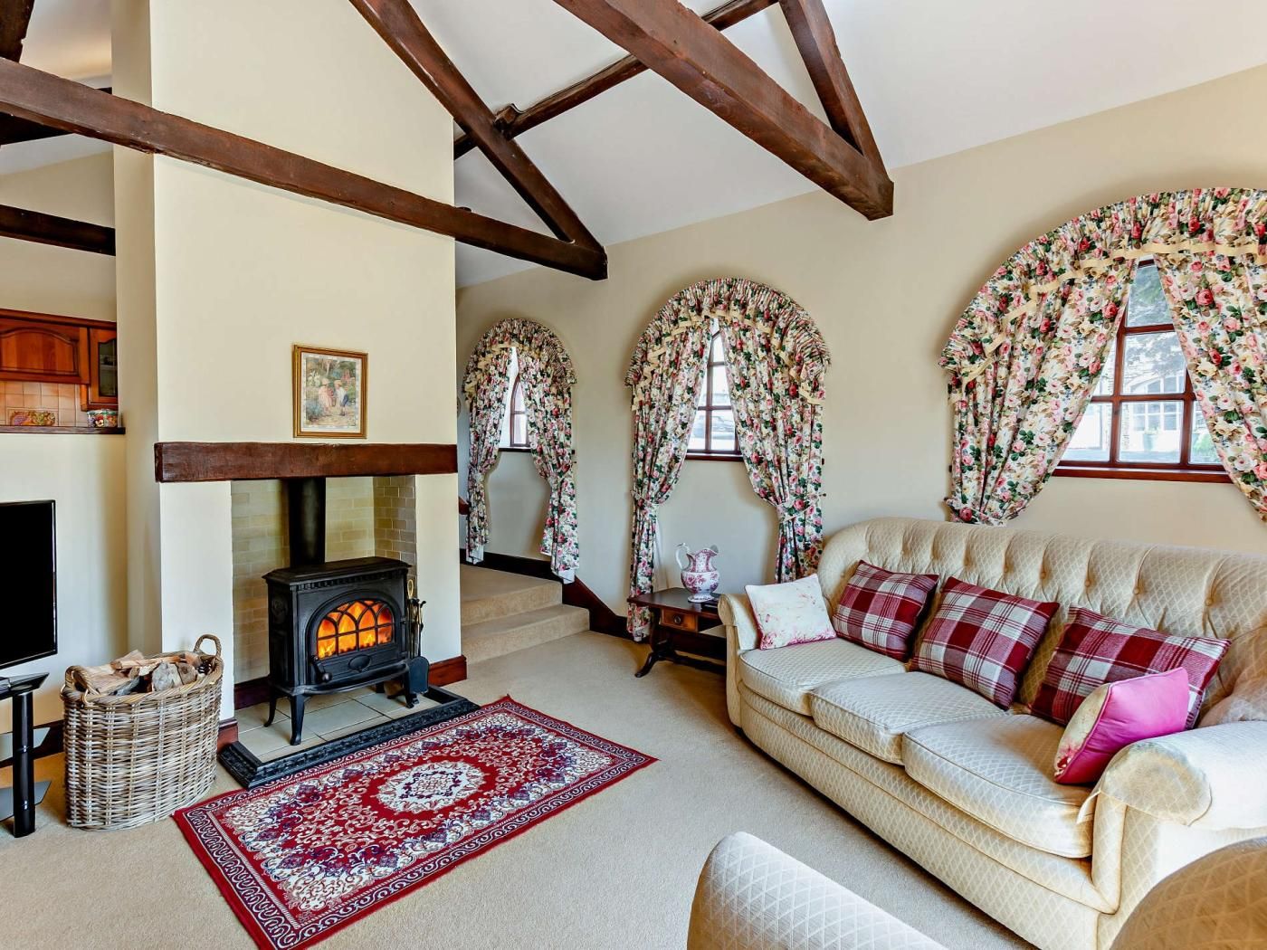 Una sala de estar acogedora con chimenea, cortinas florales, un sofá con cojines escoceses y una alfombra estampada.