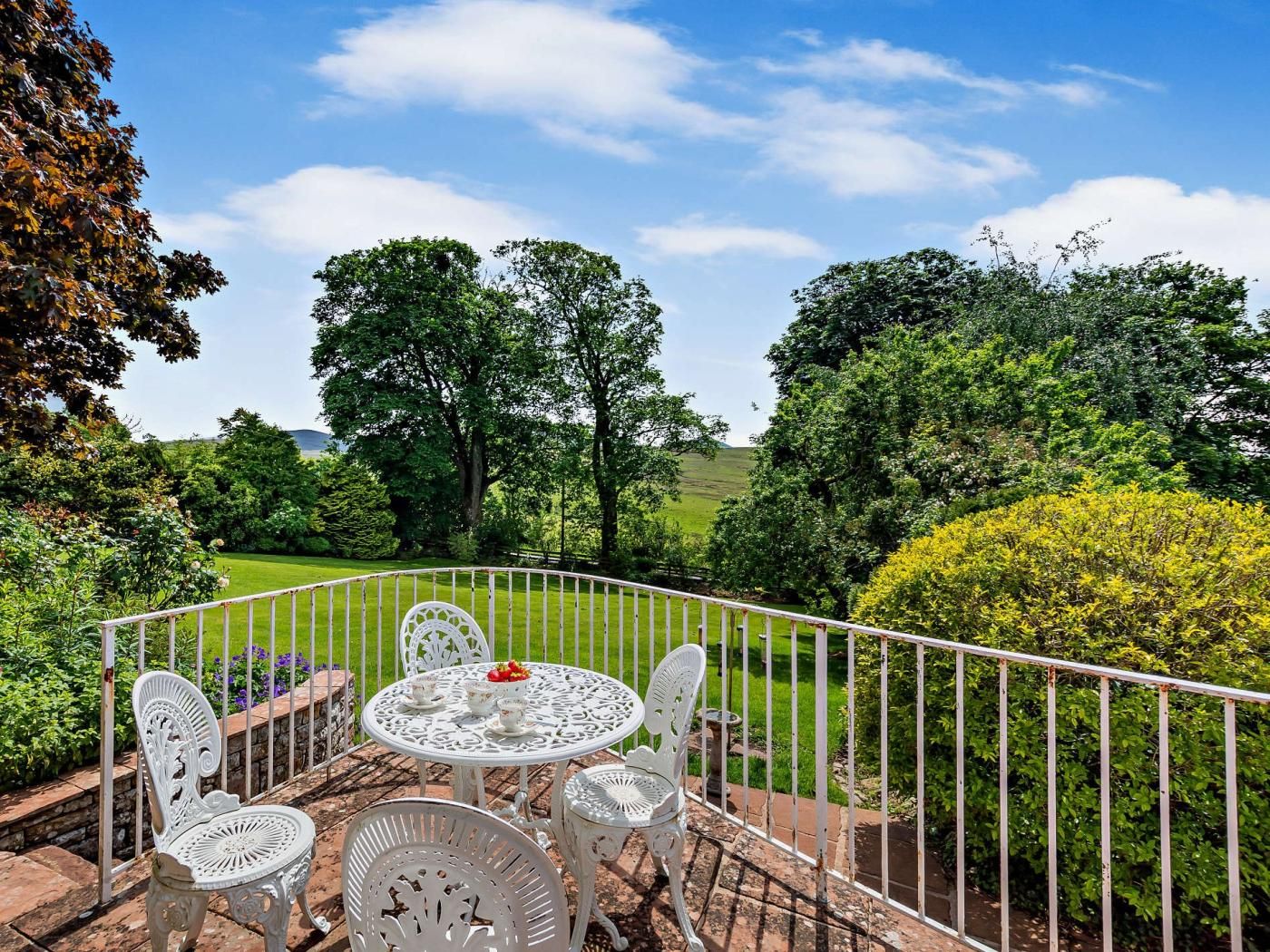 Eine Terrasse im Freien mit rundem Tisch und Stühlen, umgeben von einem weißen Metallzaun, mit Blick auf einen üppigen Garten und Bäume.