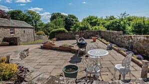 En stenbelagt terrasse med havemøbler, grill og krukker med planter.