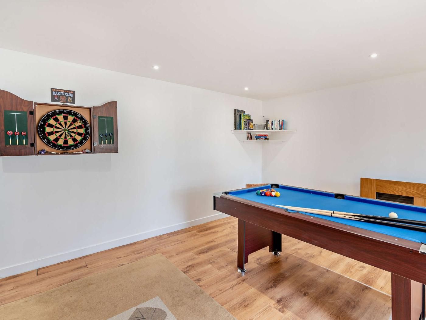 A billiards table with a blue felt surface, a dartboard, and a shelf with books and small items.