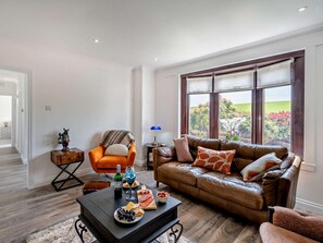 A living room with a leather sofa, a wooden coffee table with snacks, a small orange armchair, and a view of a garden through large windows.