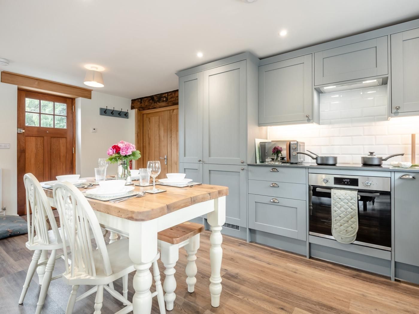 A kitchen with a wooden table set for dining, white chairs, grey cabinets, and a modern oven.