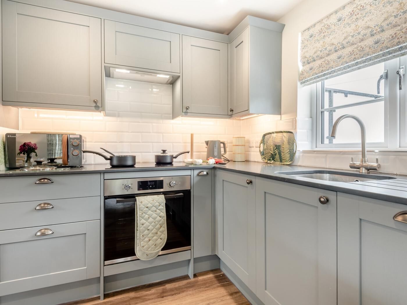 Une cuisine avec des meubles de couleur gris clair, un four encastré, une plaque de cuisson et une fenêtre agrémentée d’un rideau à motifs floraux.