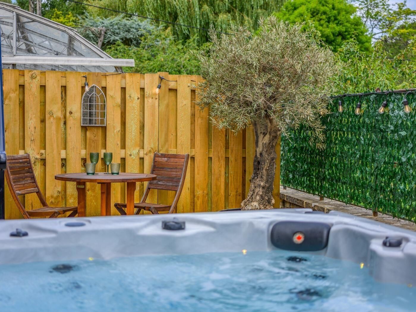 A hot tub with a wooden table and chairs, a wooden fence, and a tree.